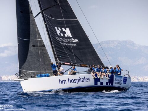 Sailing yacht with a white hull and blue sponsor logos, crew leaning on the rail as it sails across blue water with a coastal backdrop behind.