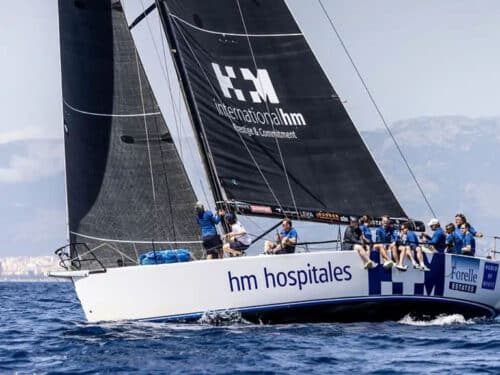 Sailing yacht with a large black mainsail marked 'HM Hospitals' and a crew in blue shirts on the deck.