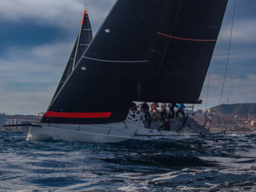 Race yacht with towering black sails cutting through waves; crew on deck near the bow as a coastal city skyline appears in the background.