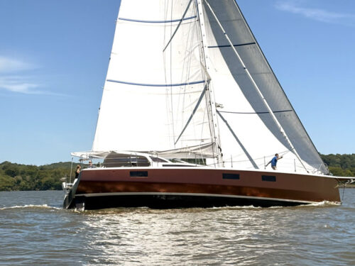 Sailing yacht with tall white sails gliding on brownish water; tree-lined shoreline under a clear blue sky.