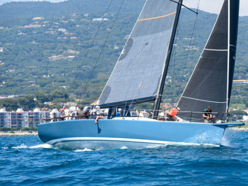 Blue racing sailboat on deep blue water near a hillside town with other sailboats in the background.