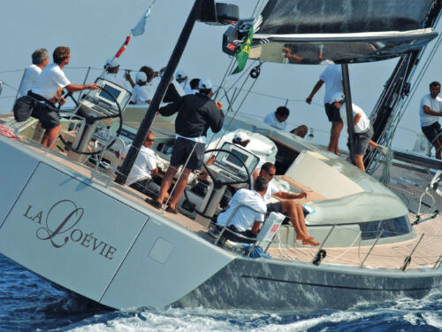 Group of people on a white luxury sailing yacht leaning to one side as it slices through blue ocean waves, with crew on deck attending to controls and sails.