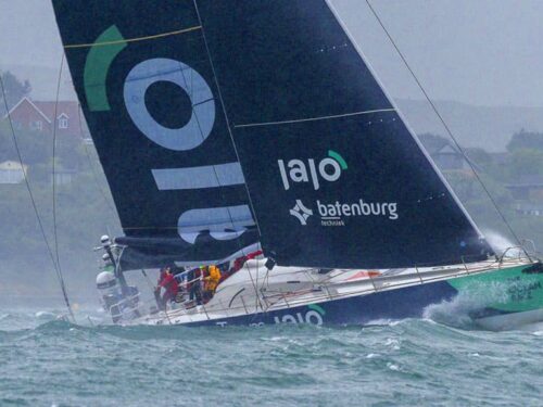 Sailing yacht edged by waves, tilted as crew in orange jackets manages the race near a residential shoreline.