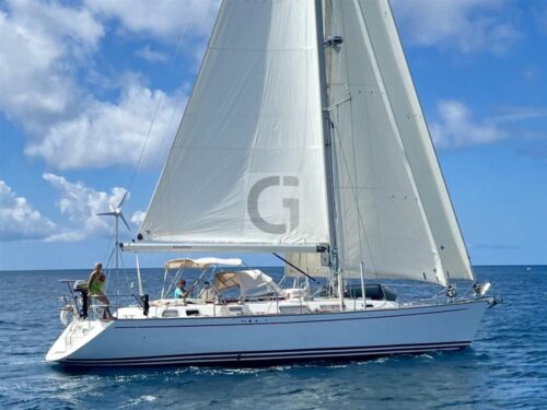 White sailing yacht with large fully raised mainsail and jib on calm blue sea, two people on deck.