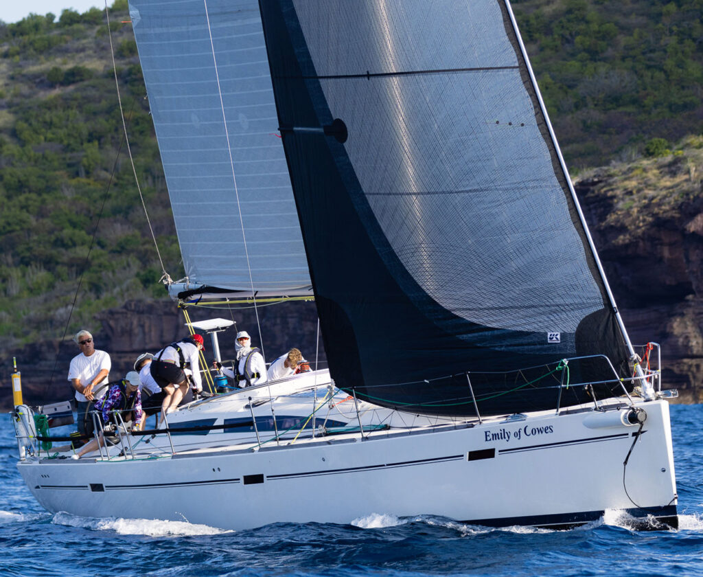 Sailing yacht named Emily of Cowes with a crew on deck, cutting through blue water near a rocky shoreline.