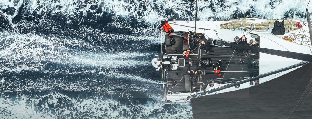 Top-down aerial view of a sailboat slicing through dark, foamy ocean with crew on deck