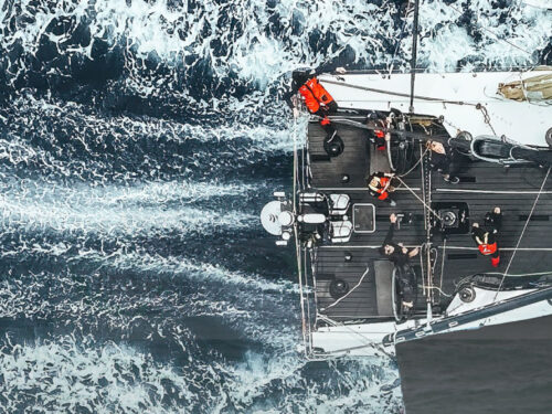 Top-down aerial view of a sailboat slicing through dark, foamy ocean with crew on deck