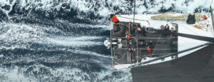 Aerial view of a sailboat cutting through blue water, white wake trailing behind as the crew works on deck.